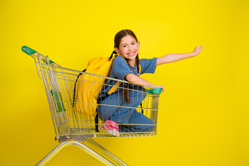 Smiling young girl with braided hair posing in a shopping cart against a vibrant yellow backdrop,...