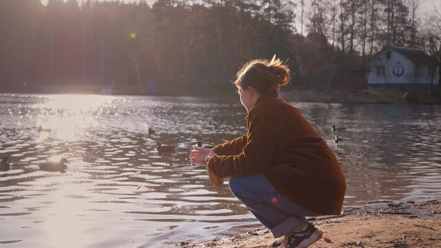 Young teenage girl dressed in casual clothes enjoying a peaceful moment by the water, squatting and feeding a group of ducks swimming in the lake, surrounded by the peaceful beauty of nature