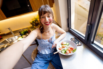 A cheerful woman takes a selfie while enjoying a healthy breakfast in a cozy kitchen. Ideal for wellness, food blogs, personal branding, or influencer content