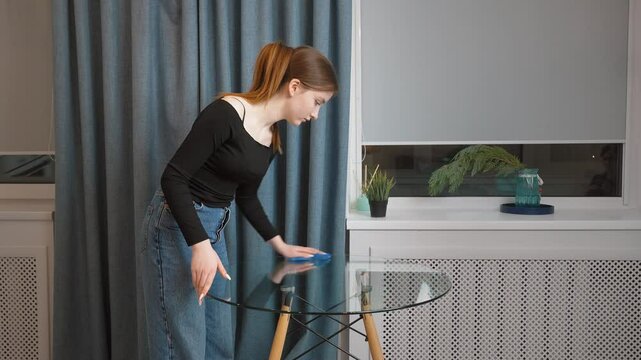 Young teen girl cleaning glass table with blue cloth, doing thorough cleaning at home with blue curtains and plant on windowsill