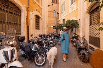 A woman in a blue coat walks her white dog through a narrow Roman alley filled with parked...
