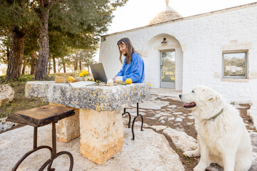 A woman works on her laptop at a rustic stone table in front of a trullo house with her dog nearby....