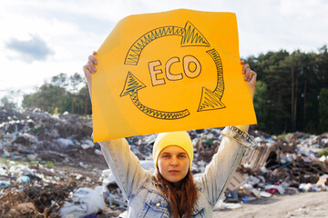 A young woman raises a yellow ECO recycling symbol sign at a waste site, advocating for...