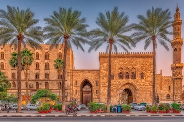 Naklejka premium Palm trees line the street in Cairo, showcasing beautiful historic buildings at dusk. The architecture features intricate designs, highlighting the rich culture and history of the area.