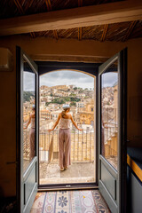 Woman standing on a balcony with panoramic view over the baroque rooftops and historic church of a hilltop Sicilian town. Reflection in glass door, soft light and serene atmosphere