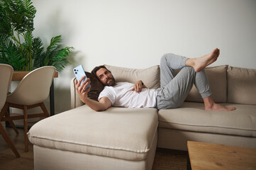 Obraz premium Man Relaxing on Sofa Holding Smartphone in Cozy Minimalist Living Room Setting