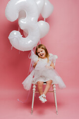 little girl with balloons and the number three on a pink background celebrates her birthday in the studio