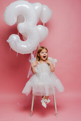 little girl with balloons and the number three on a pink background celebrates her birthday in the studio