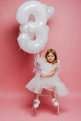 little girl with balloons and the number three on a pink background celebrates her birthday in the studio