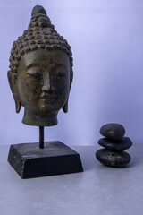 Serene Buddha head statue beside stacked black stones in a calm, minimalist setting
