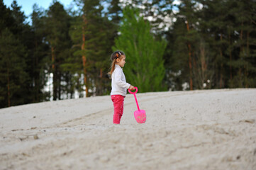 Cute child girl walking with shovel in her hand on the sand on nature background in the spring. Happy childhood, space for text