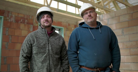 Two construction workers posing indoors at building site, men wearing safety helmets stand together in unfinished structure, team camaraderie and job pride