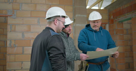 Construction workers discussing building plans on-site, team collaboration, three men in safety helmets reviewing architectural blueprints in an unfinished brick wall interior