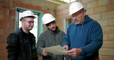 Construction workers discussing project details, three men in safety helmets reviewing blueprints and collaborating on-site in an unfinished brick structure, indoor team planning