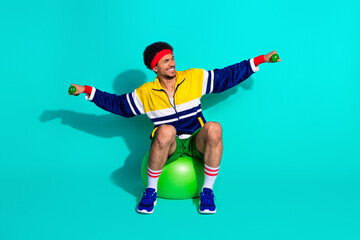 Young man sitting on a gym ball in fitness attire while exercising with dumbbells against a vibrant turquoise background.