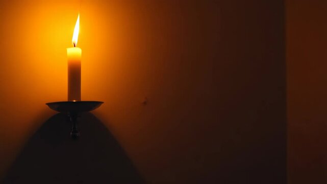 Single burning wax candle mounted on ornate metal candle holder casting shadows against a neutral tan colored wall in dimly lit room.