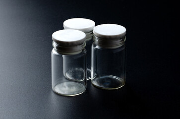Three small empty transparent glass jars with white plastic screw caps arranged on a dark textured surface in studio lighting, ideal for packaging, storage or pharmaceutical concepts