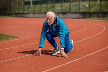 An old sportsman kneeling in start position and preparing for running on stadium.