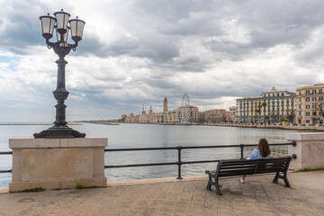 Panorama of Bari, Puglia or Apulia, southern Italy with Mediterranean Sea, street lamps, ferris wheel and a girl or woman sitting on the bench on the promenade with storm clouds