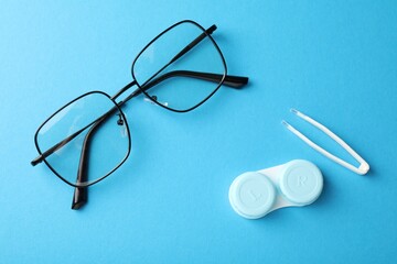 Contact lens case, tweezers and glasses on light blue background, flat lay
