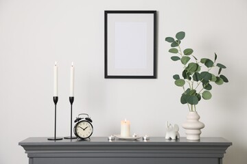 Stylish candlesticks with burning candles, other decor and alarm clock on fireplace near white wall...