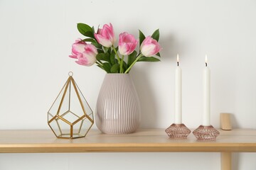 Elegant holders with burning candles, tulips in vase and other decor on wooden table near white wall