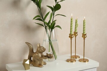 Elegant candlesticks with burning candles, branches in vase and other decor on dresser near beige...