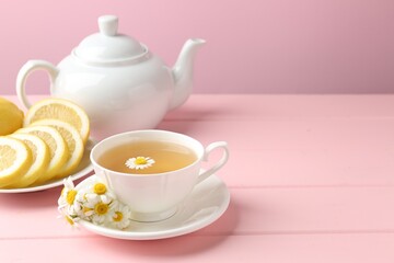 Delicious chamomile tea in cup, fresh flowers and lemon on pink wooden table, closeup. Space for text