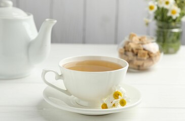 Delicious chamomile tea in cup, fresh flowers and brown sugar on white wooden table, closeup