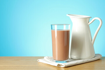 Tasty chocolate milk in glass and jug on wooden table against light blue background, space for text