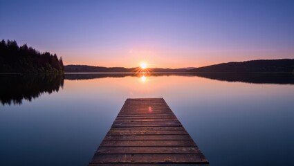 Fototapeta premium Wooden pier extending into a tranquil lake at sunset with forest view