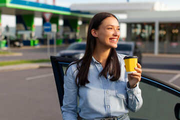Happy woman driver having coffee break standing near her car on the gas station, making stop during...