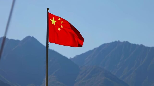 Red flag of China with yellow stars waving dynamically in wind against a backdrop of majestic mountains and clear blue sky.