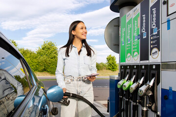 Best fuel price. Happy woman refueling her automobile tank with bio fuel and holding cellphone,...