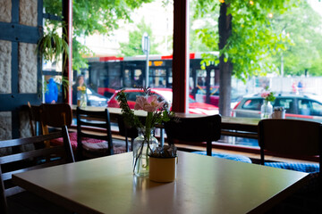 Cozy urban café interior with flowers on table and blurred street view through large window featuring bus, cars, and pedestrians, evoking relaxed atmosphere in vibrant city neighborhood.