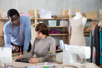 Two interested fashion designers, man and woman, collaborating in sewing studio, using tablet to create and refine new garment design at pattern table, discussing digital sketches or client references