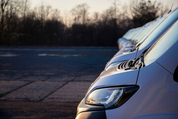 Side and front of a panel van in a long row of identical white vans ready for customer delivery. Headlight, hood, fender, bumper. A line of vans in a logistics parking lot. © Prokop