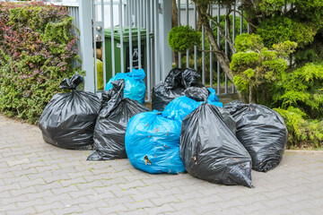 Black and Blue Garbage Bags Gathered Near Decorative Garden Plants and White Fence