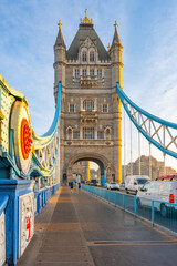 Obraz premium Soft morning light casts a warm glow on Tower Bridge. Pedestrians walk along the pathway while vehicles pass, showcasing the iconic structure against a clear sky.