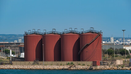 Four large, red oil storage tanks stand prominently in a white bay, with a cityscape visible in the background under a clear blue sky