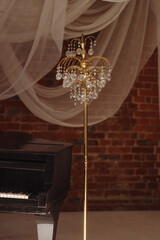 A golden crystal floor lamp stands next to an old black upright piano, set against a brick wall with draped white fabric. Concept vintage style, music, or artistic interiors.