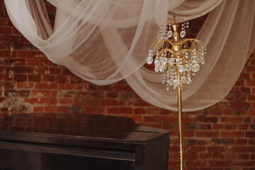 A golden crystal floor lamp stands next to an old black upright piano, set against a brick wall...