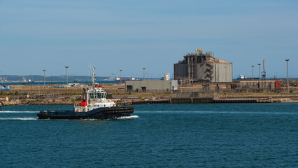 Fototapeta premium A tugboat moves past a large, concrete LNG storage tank in a busy industrial port