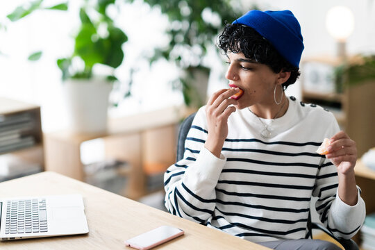 A person in a blue beanie and striped shirt is eating a tangerine at a desk. They are taking a break from work to enjoy a healthy snack.