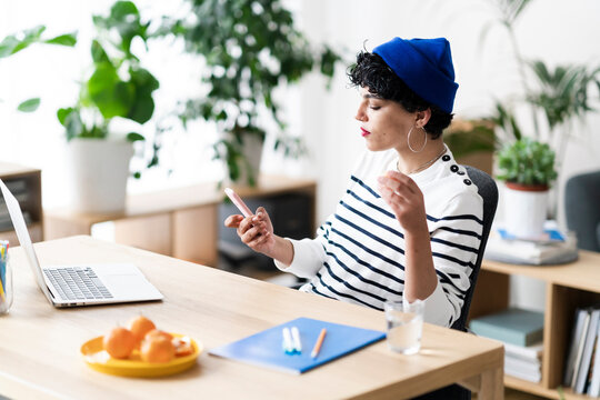 A woman in a blue beanie is taking a break from her laptop to check her phone and eat an orange. She's likely catching up on messages or social media.