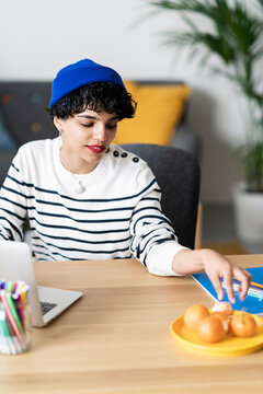 A woman is working from home at her desk. She's reaching for her notebook, perhaps to jot down ideas or notes while she works on her laptop.