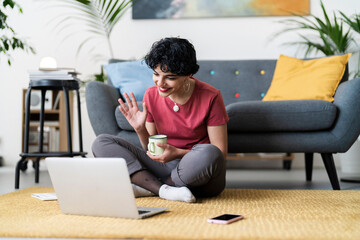 A woman is video chatting on her laptop while sitting on the floor with a mug. She's connecting with someone remotely, likely for work or to catch up with friends/family.