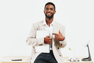 Confident smiling man in beige blazer holding laptop and pointing at it, looking at camera in...