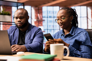 Obraz premium African American freelancers at home collaborating on a research project, man using online reports and analytics via laptop. Young woman manages web correspondence on mobile phone.