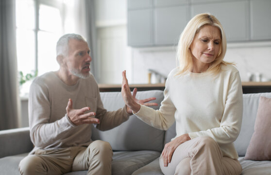 A senior man and a woman sit on a couch, engaged in a heated discussion. The woman appears upset and is signaling for the man to stop talking, indicating tension in their relationship.